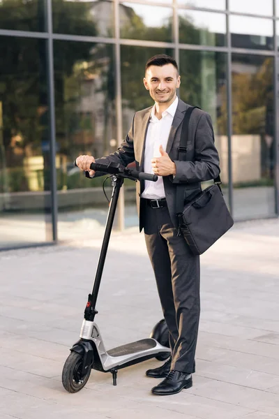 İş binasının önünde elektrikli scooterıyla yakışıklı bir iş adamı. Zarif takım elbiseli genç işadamı, dışarıda duruyor, neşeyle gülümsüyor ve başparmağını gösteriyor. — Stok fotoğraf