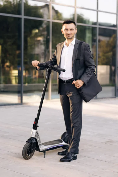 Şehrin önünde elinde elektrikli scooterla kameraya bakan bir iş adamı. Modern kentsel alternatif ulaşım. Çevre dostu ulaşım. Ekoloji ve kentsel yaşam tarzı — Stok fotoğraf