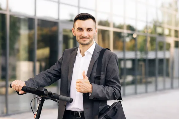 Zarif takım elbiseli genç bir işadamı dışarıda, neşeyle gülümsüyor ve başparmağını kaldırıyor. İş binasının önünde elektrikli scooterıyla yakışıklı bir iş adamı.. — Stok fotoğraf