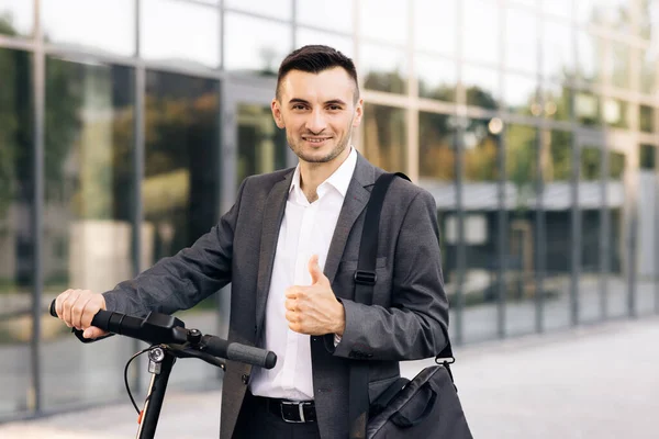 Zarif takım elbiseli genç bir işadamı dışarıda, neşeyle gülümsüyor ve başparmağını kaldırıyor. İş binasının önünde elektrikli scooterıyla yakışıklı bir iş adamı. — Stok fotoğraf