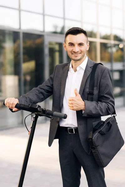 İş binasının önünde elektrikli scooterıyla yakışıklı bir iş adamı. Zarif takım elbiseli genç işadamı, dışarıda duruyor, neşeyle gülümsüyor ve başparmağını gösteriyor.. — Stok fotoğraf