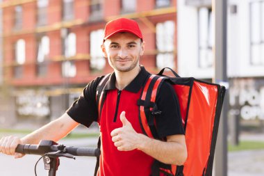 Portre gülümseyen Amerikan kurye yiyecek teslimatı termal sırt çantasıyla ayakta duruyor, elektrikli scooter kameraya bakıyor ve başparmağını kaldırıyor. Olumlu teslimatçı işçi online sipariş istemcisi teslim.