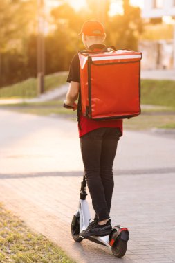 Tanımlanamayan genç kurye e-scooterıyla geziyor. Erkek paket teslimatçısı, gün batımında sokağın aşağısındaki kırmızı sırt çantalı, boş bir kaldırımdan elektrikli scooter kullanıyor.