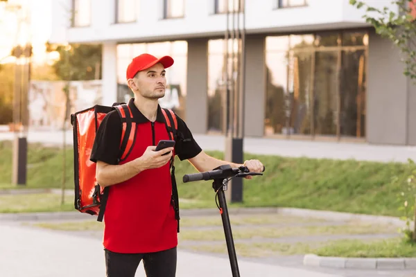 Kırmızı termal sırt çantalı erkek kurye yiyecek teslimatı. Elektrikli scooterlı caddede yürüyor. Akıllı telefon navigasyonu kullanıyor. Teslimatçı işçi gün batımında müşterileri online sipariş ile teslim ediyor.. — Stok fotoğraf
