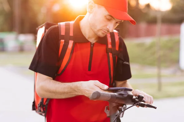 Kırmızı termal sırt çantalı kurye yemek teslimatı. Elektrikli scooterlı caddede yürüyor. Akıllı telefon navigasyonu kullanıyor. Teslimatçı işçi çevrimiçi sipariş müşterisini teslim ediyor. — Stok fotoğraf