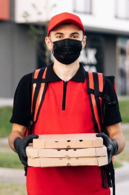 Karton kutularda pizza taşıyan, koruyucu maske ve eldiven takan kırmızı sırt çantalı kurye adam portresi. Mükemmel atış. Teslimat hizmetleri