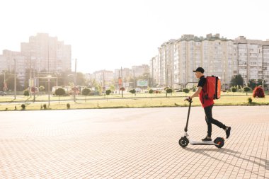 Asyalı kurye yemek teslimatı. Kırmızı termal sırt çantalı. Elektrikli scooter ile sokakta geziyor. Kırmızı üniformalı teslimatçı işçi. Mükemmel atış.