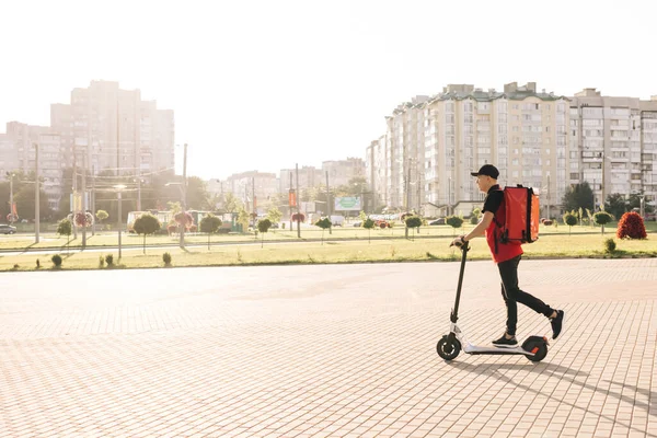 Asyalı kurye yemek teslimatı. Kırmızı termal sırt çantalı. Elektrikli scooter ile sokakta geziyor. Kırmızı üniformalı teslimatçı işçi. Mükemmel atış. — Stok fotoğraf