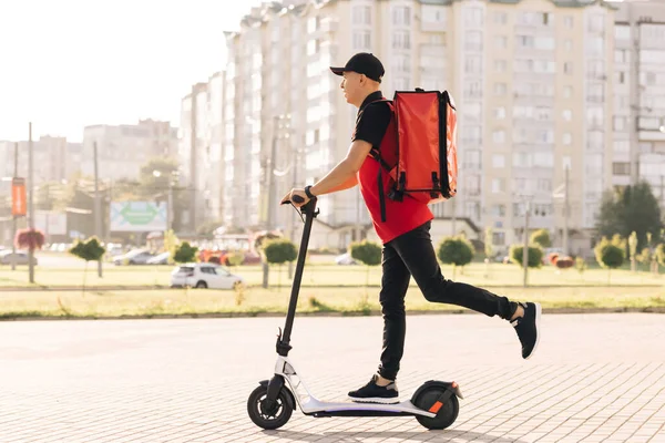Kırmızı termal sırt çantalı erkek kurye yemek teslimatı elektrikli scooter ile caddeye çıkıp müşteriye sipariş veriyor. Kırmızı üniformalı teslimatçı işçi. Mükemmel atış. — Stok fotoğraf