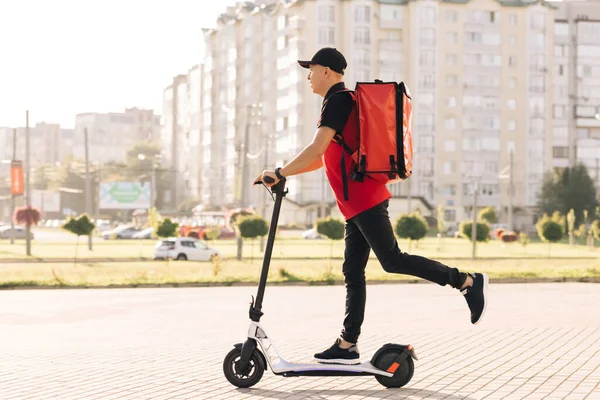 Hombre mensajero entrega de alimentos con mochila térmica roja paseos por la calle en un scooter eléctrico entregar cliente de pedidos en línea. Trabajador de reparto con uniforme rojo. Excelente entrega — Foto de Stock