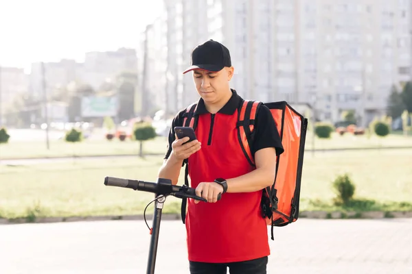 Entrega de alimentos mensageiro masculino com mochila térmica vermelha olha em torno da rua com scooter elétrico usa smartphone navegar. Trabalhador entregador empregado entregar cliente cliente ordem on-line — Fotografia de Stock