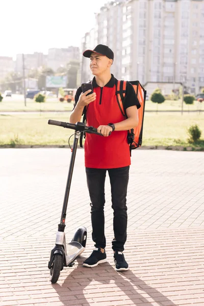 Mannelijke koerier man is op zoek naar mobiele telefoon om het adres van voedsel te leveren van een restaurant. Voedsel bezorger navigeert in de telefoon in de stad met elektrische scooter. Milieuvriendelijk vervoer — Stockfoto