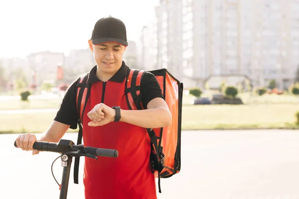 Positive deliveryman worker employee deliver online order client customer. Smiling man courier food delivery with red thermal backpack walks street with electric scooter