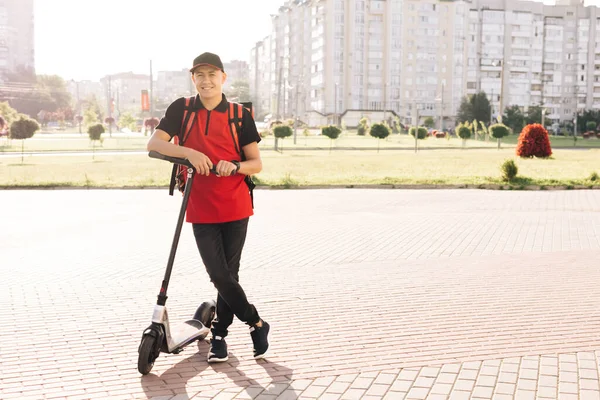 Portret van bezorger staande met elektrische scooter met rode thermische rugzak kijkend naar camera. E-Scooter rijder eco transport. Modern stedelijk alternatief vervoer. Uitstekende levering — Stockfoto