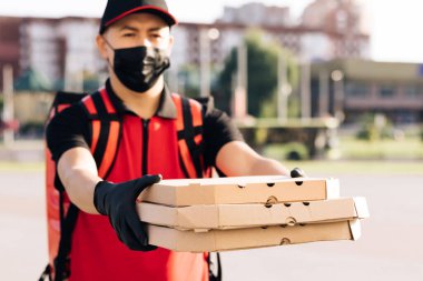 Eldivenli ve tıbbi maske takmış yakışıklı beyaz bir teslimatçının elinde karton kutular tutarken çekilmiş portresi. Erkek kurye pizza veriyor.