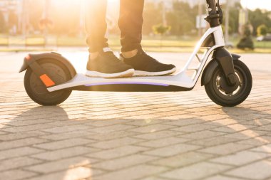 Foot 'u kapatın kurye yemek teslimatı elektrikli scooter' la yolda gidiyor günbatımında internetten sipariş veren müşteriyi teslim ediyor. Siyah spor ayakkabılı genç adam elektrikli mobilete biniyor.