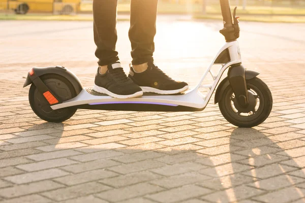 Disparo en las piernas joven hipster hombre en zapatillas negras paseo en scooter móvil eléctrico al atardecer. Hombre hipster montando un scooter eléctrico. Transporte ecológico. Transporte eléctrico de conducción rápida — Foto de Stock