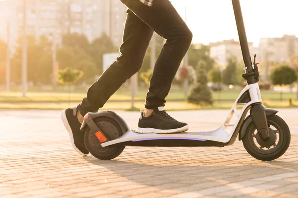 Disparo en las piernas joven en zapatillas negras paseo en scooter móvil eléctrico. Close up feet man courier food delivery paseos por la calle en scooter eléctrico entregar pedido en línea cliente al atardecer — Foto de Stock