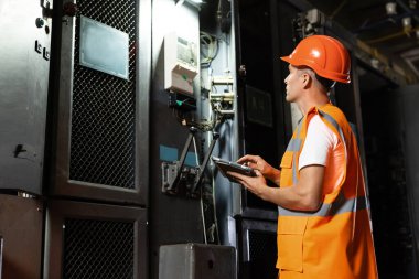 Sert şapka ve yelek giyen bir elektrik istasyonu mühendisi rafa doğru yürür ve dijital tablet kullanarak elektrik kontrol odasındaki elektrik güç sistemini inceler. Sanayi, fabrika, enerji kavramı