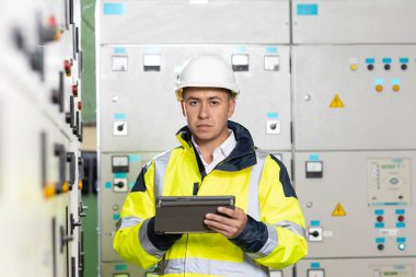 Asyalı elektrik mühendisi kontrol odasındaki elektrik sistemini izlemek için tablet tutuyor. Üretimde fabrika elektriği üretmek için ekran sistemini kontrol eden teknisyen teftişi