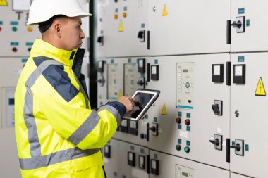 Asyalı elektrik mühendisi kontrol odasındaki elektrik sistemini izlemek için tablet tutuyor. Üretimde fabrika elektriği üretmek için ekran sistemini kontrol eden teknisyen teftişi