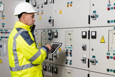 Asyalı elektrik mühendisi kontrol odasındaki elektrik sistemini izlemek için tablet tutuyor. Üretimde fabrika elektriği üretmek için ekran sistemini kontrol eden teknisyen teftişi