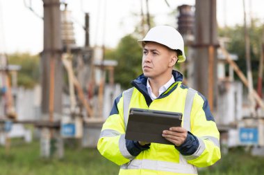 Yüksek voltajlı elektrik pilonu olan elektrik mühendisi. Özel elbiseli bir enerji mühendisi dijital tablet bilgisayarındaki elektrik sensörlerinden elde edilen verileri kullanarak güç hattını kontrol ediyor.