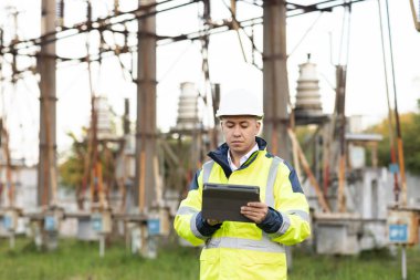 Özel elbiseli enerji mühendisi dijital tablet bilgisayarındaki elektrik sensörlerinden elde edilen verileri kullanarak bir elektrik hattını inceliyor. Yüksek voltajlı elektrik direğine sahip elektrik mühendisi.