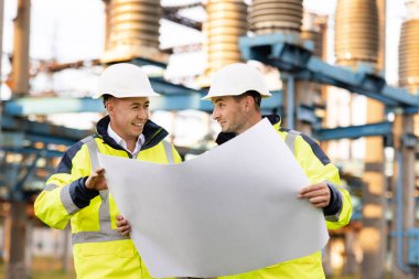 İki mühendis elektrik nakil hatlarının önünde yenilenebilir enerji geliştirme üzerinde çalışıyorlar. Özel kıyafetli mühendisler yüksek voltajlı elektrik hattının yakınındaki kağıt üzerinde bir çizimi tartıştılar.