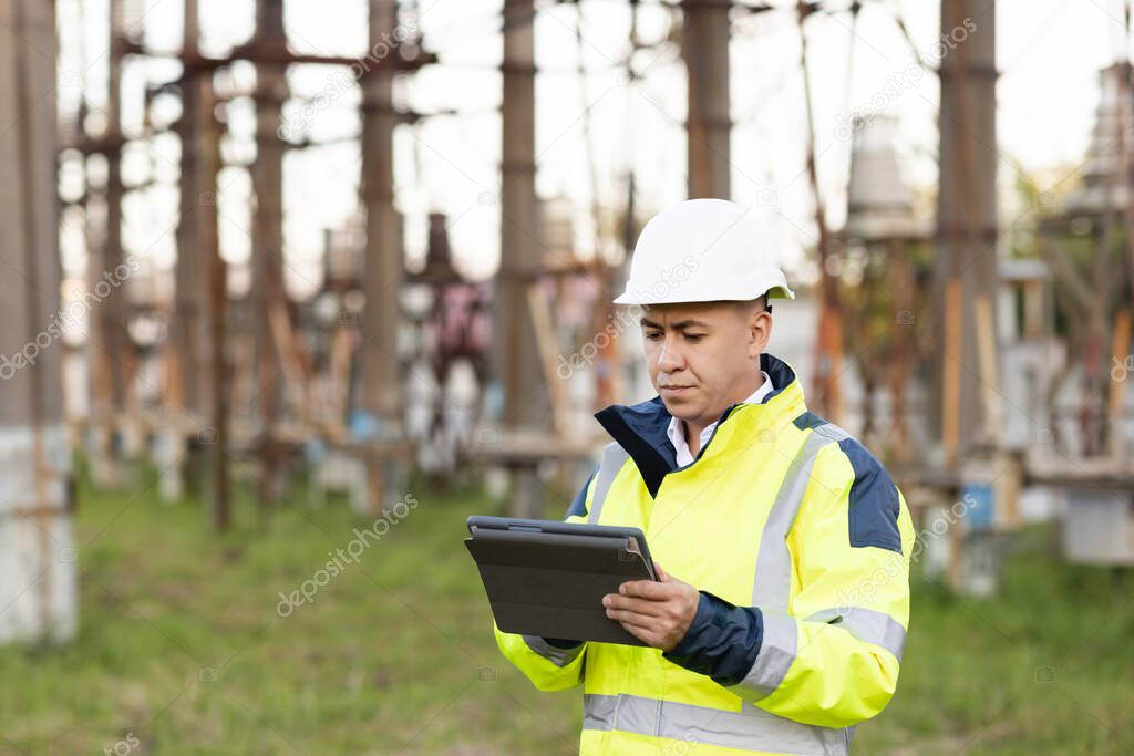 An energy engineer man in special clothes inspects a power line using data from electric sensors on a digital tablet computer. Electrical engineer with high voltage electricity pylon