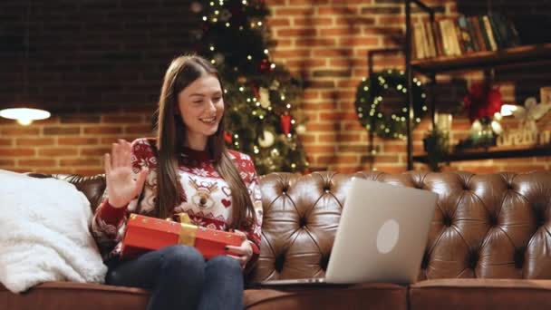 Excité heureux caucasien fille porte santa chapeau détient boîte cadeau de Noël, ouvre cadeau de Noël sur ordinateur vidéoconférence chat de réunion virtuelle avec la famille, ami de distance sociale se trouve sur le canapé à la maison — Video