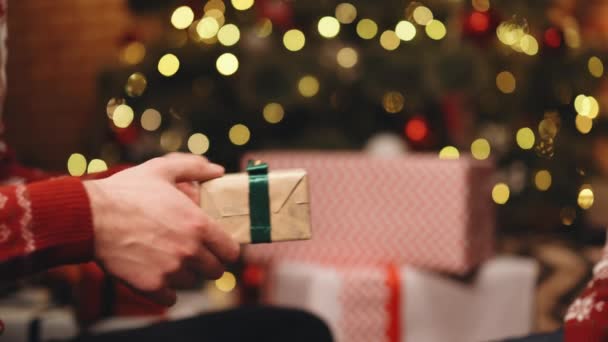 Gros plan mains de l'homme donnant boîte de fête avec cadeau de Noël à la femme assise à la table de fête pendant la fête de famille de vacances, décoration arbre de Noël et lumières de célébration, foyer sélectif — Video
