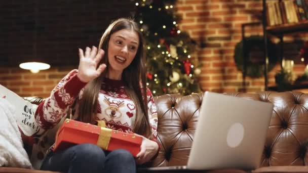 快乐的年轻的高加索女士坐在沙发上聊天，在笔记本电脑上的视频会议应用程序，向网络摄像头展示礼品盒。兴奋的千禧年女士用pc向好朋友问候新年。圣诞老人的礼物 — 图库视频影像