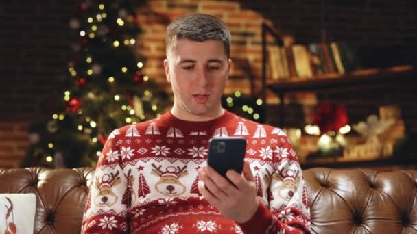 Portret szczęśliwego białego mężczyzny uśmiechającego się z zaskoczoną miną stukającego w smartfona czując podniecenie i wygrywając świąteczną loterię patrząc w kamerę. Wesołych Świąt Bożego Narodzenia koncepcja — Wideo stockowe