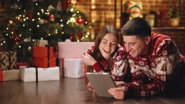 タブレットを使用して祝福メリークリスマスの親戚や友人とクリスマスツリー輝くガーランドテキストメッセージsmsで飾られた家のホールで寝そべってカップル。オンライン、販売、近代的な技術の過剰使用の概念を購入する — ストック動画