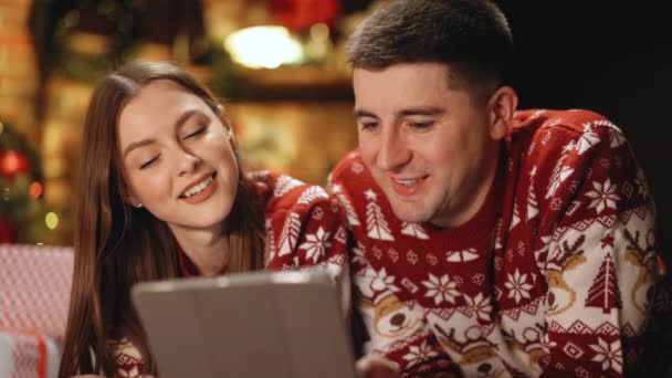 Feliz pareja caucásica acostada en casa cerca del fondo del árbol de Navidad. Hermosa mujer con el pelo largo sostiene tableta digital y emocionalmente cuenta historias divertidas — Vídeos de Stock