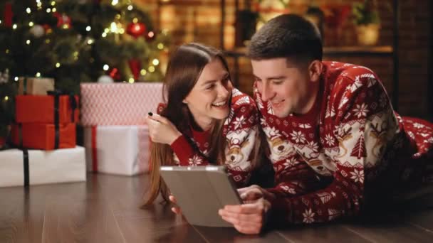 Кавказька сім'я вибирає різдвяний подарунок онлайн на планшеті, лежачи в затишному будинку. Чоловік і дружина шукають подарунок від xmas на планшетному комп "ютері. Дух Нового року. Святкування свят — стокове відео