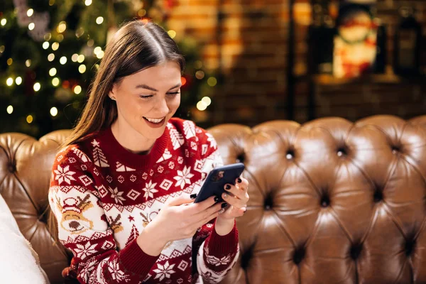 Akıllı telefon kullanan neşeli güzel bir kadın, çevrimiçi oyunlar oynuyor, sosyal ağda iletişim kuruyor, arkadaşlarına ya da ailesine Noel tebriklerini iletiyor evdeki Noel ağacının yanındaki kanepede dinleniyor.