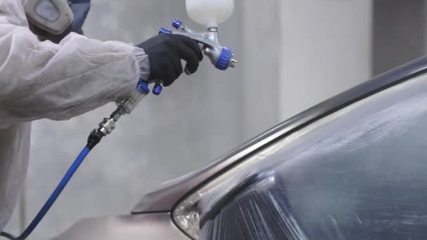 Egy férfi, aki autószervizben dolgozik, egyenruhában, és maszkot visel, autóval, légmentes szórópisztollyal a fülkében. Járműalkatrészek festése autószervizműhelyben. — Stock videók