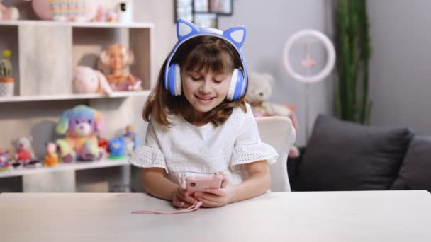 Симпатична маленька дівчинка в навушниках використовує смішний додаток для редагування на смартфоні, насолоджуючись крутим відео або музичним контентом у соціальній мережі, граючи в онлайн-ігри, віддалено спілкуючись — стокове відео