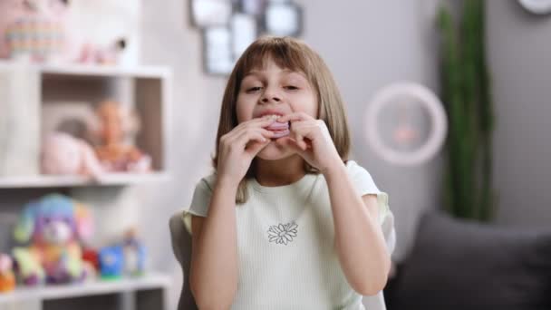 Aantrekkelijk brunette school meisje op een thuis achtergrond, likt haar lippen, kijkt naar camera eten een violette macaron, sluit haar ogen met plezier — Stockvideo