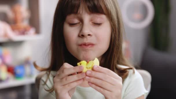 Приваблива брюнетка школярка на домашньому тлі, лиже губи, їсть одного жовтого макарона, із задоволенням закриває очі. Десерт, солодкий зуб, гастрономічний. Відчуваю себе добре, смачно — стокове відео