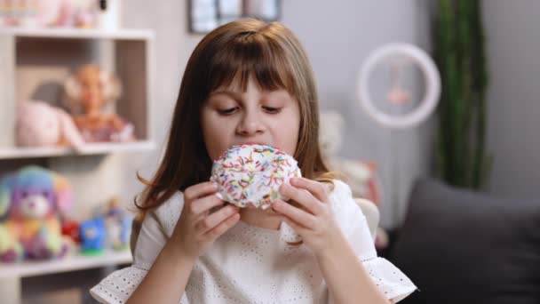 Grappig klein meisje dat lekkere donut eet op haar achtergrond. Schattige vrouw die donut eet. Schattig kind meisje bijten stuk donut terwijl zitten aan tafel thuis — Stockvideo