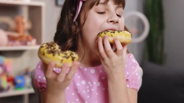 Mooi meisje eten donut genieten van zoete heerlijke smaak van glazuur en hagelslag glimlachen met tevredenheid. Schoolmeisje eet twee donuts tegelijk. Zoete verslaving. — Stockvideo