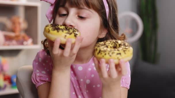 Fechar menina da escola comer dois donuts enquanto sentado à mesa em casa. Retrato de menina doce escolher entre dois donuts dentro de casa. Engraçado adolescente menina se divertindo com donuts coloridos em casa moderna — Vídeo de Stock