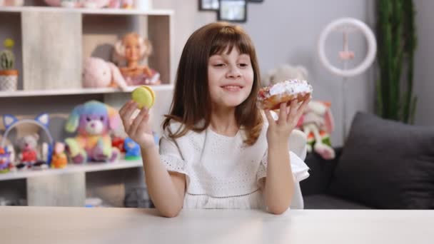 Heerlijk vrolijk schoolmeisje kan niet kiezen tussen een witte donut en gele verse makaken. Grappig tienermeisje die plezier heeft met donut en macaron in het moderne huis. Zoetekauw. Ongezond voedselconcept — Stockvideo