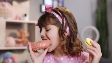 Evdeki masada otururken tatlı bir kız çöreğin bir parçasını ısırıyor. Beyaz liseli kız donut yiyor ve sarı makaron yiyor ve kameraya bakıyor. Tatlı bağımlılık konsepti