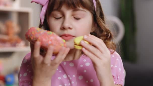 Gros plan d'une jolie écolière sur fond de maison, se lèche les lèvres, renifle le beignet et le macaron jaune, ferme les yeux avec plaisir. Dessert, sucré, gourmand. Je me sens bien, délicieux — Video