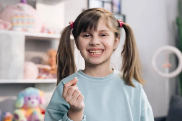 Portrait of cute little child girl is rejoices and showing her lost milk tooth and smiling to camera of toothless mouth while she standing in bedroom. Happy girl holding her fallen tooth in hand. — Stock Photo, Image