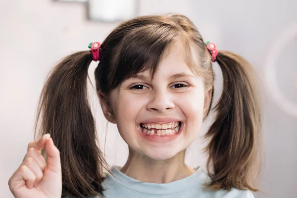 Portrait of cute little child girl is rejoices and showing her lost milk tooth and smiling to camera of toothless mouth while she standing in bedroom. Happy girl holding her fallen tooth in hand. — Stock Photo, Image